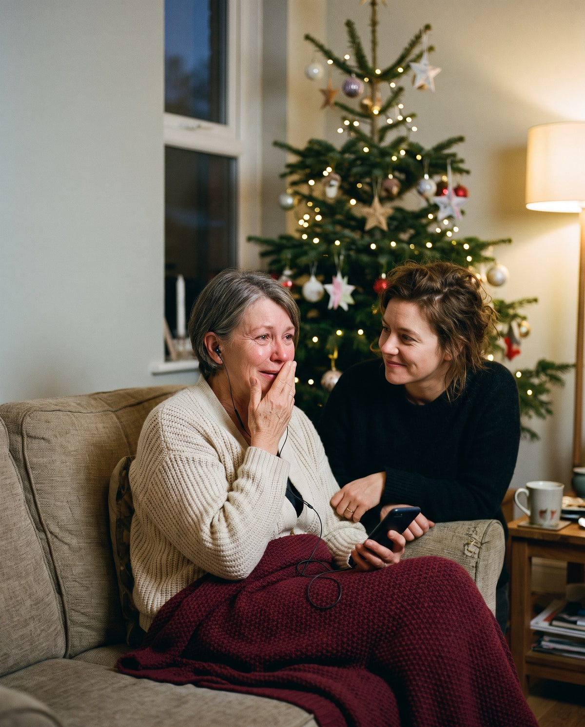 Gerührte Mutter hört mit Kopfhörern ein Lied auf dem Handy, die Tochter sitzt lächelnd neben ihr, im Hintergrund der Weihnachtsbaum