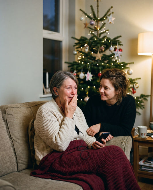 Gerührte Mutter hört mit Kopfhörern ein Lied auf dem Handy, die Tochter sitzt lächelnd neben ihr, im Hintergrund der Weihnachtsbaum